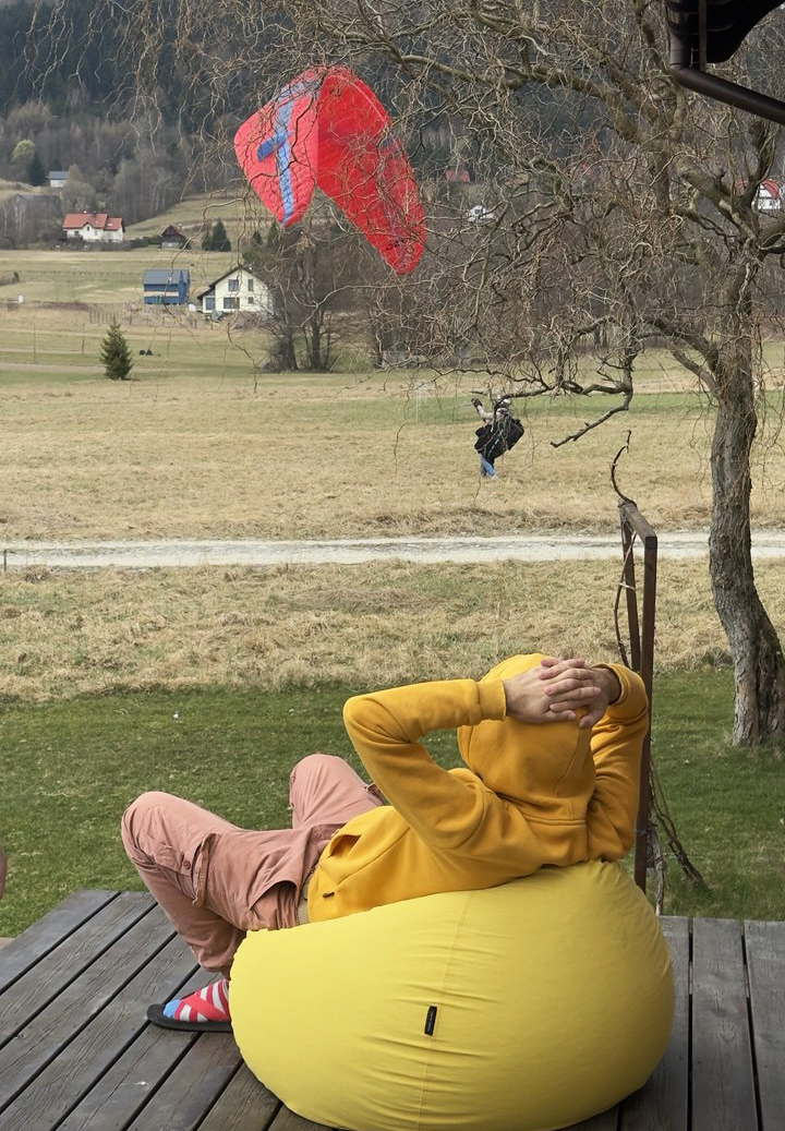 Na pierwszym planie mężczyzna siedzący na pufie obserwujący pilota paralotni trenującego ground-handling